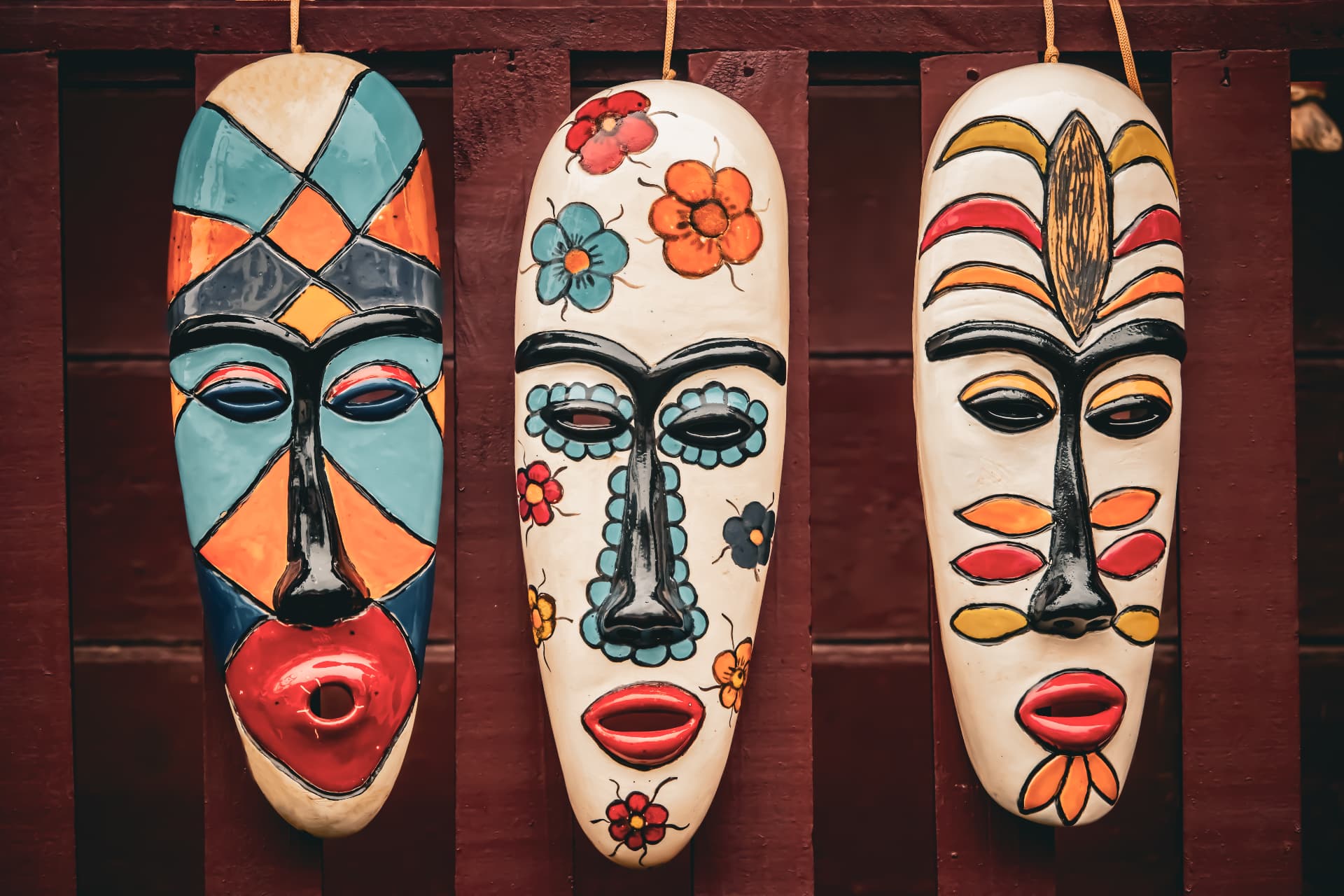 Máscaras africanas decorativas colgadas en una pared de madera, elaboradas artesanalmente y pintadas a mano con colores intensos, patrones geométricos y motivos florales de inspiración cultural africana