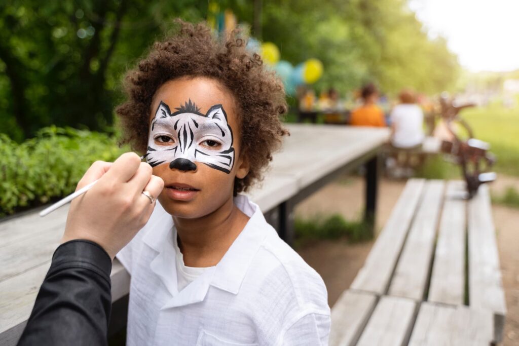 Niño en actividad con máscaras pintadas, con la cara decorada como un tigre mientras un adulto le aplica pintura facial durante una actividad infantil al aire libre, en un entorno natural con mesas de madera y ambiente festivo