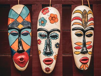 Máscaras africanas decorativas colgadas en una pared de madera, elaboradas artesanalmente y pintadas a mano con colores intensos, patrones geométricos y motivos florales de inspiración cultural africana